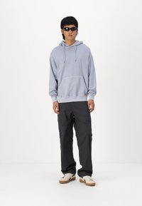 Lys grå hættetrøje med en frontlomme, kombineret med mørke cargo-bukser og hvide sneakers med brune såler. Firkantede sorte solbriller.