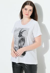 Světle šedé bavlněné tričko s černobílým grafickým motivem kozy na přední straně, kulatým výstřihem a krátkými rukávy. Nosené s černými koženými kalhotami.