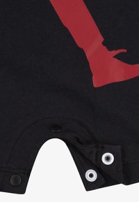 Detailní záběr na černou tkaninu se grafickým motivem červené boty a černobílými patentkami na okraji oděvu.