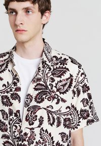 Kurzarmhemd mit cremiger Basis, das detaillierte burgunderrote Paisley- und Blumenmuster sowie ein klassisches Kragen-Design aufweist.