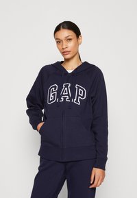 Námornícky zipsový mikina s bielym logom "GAP", kapucňou, prednými vreckami a rebrovanými manžetami. Mäkký materiál a voľný strih. Skombinované s ladiacimi nohavicami.