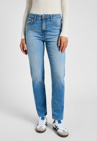 Jeans de mezclilla de color azul claro, con un corte recto, cinco bolsillos y cierre de botón metálico. Combinados con zapatillas blancas.