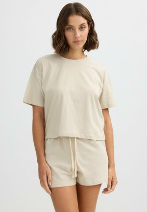 Mujer con cabello castaño hasta los hombros que lleva una camiseta beige de manga corta y pantalones cortos a juego con cordón, de pie frente a un fondo liso.
