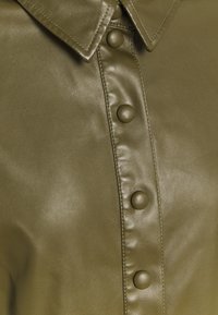 Olivgrünes Ledershirt mit einem strukturierten Kragen, großen passenden Knöpfen entlang der Vorderseite und einer glatten, strukturierten Oberfläche.