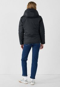 Giacca a vento nera con cappuccio, superficie texturizzata e silhouette leggermente aderente. Abbinata a jeans blu e sneakers bianche.