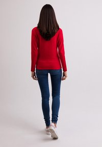 Rotes Langarmoberteil kombiniert mit blauen, eng anliegenden Jeans und weißen Sneakers. Das Oberteil hat ein schlichtes Design mit einer taillierten Silhouette.