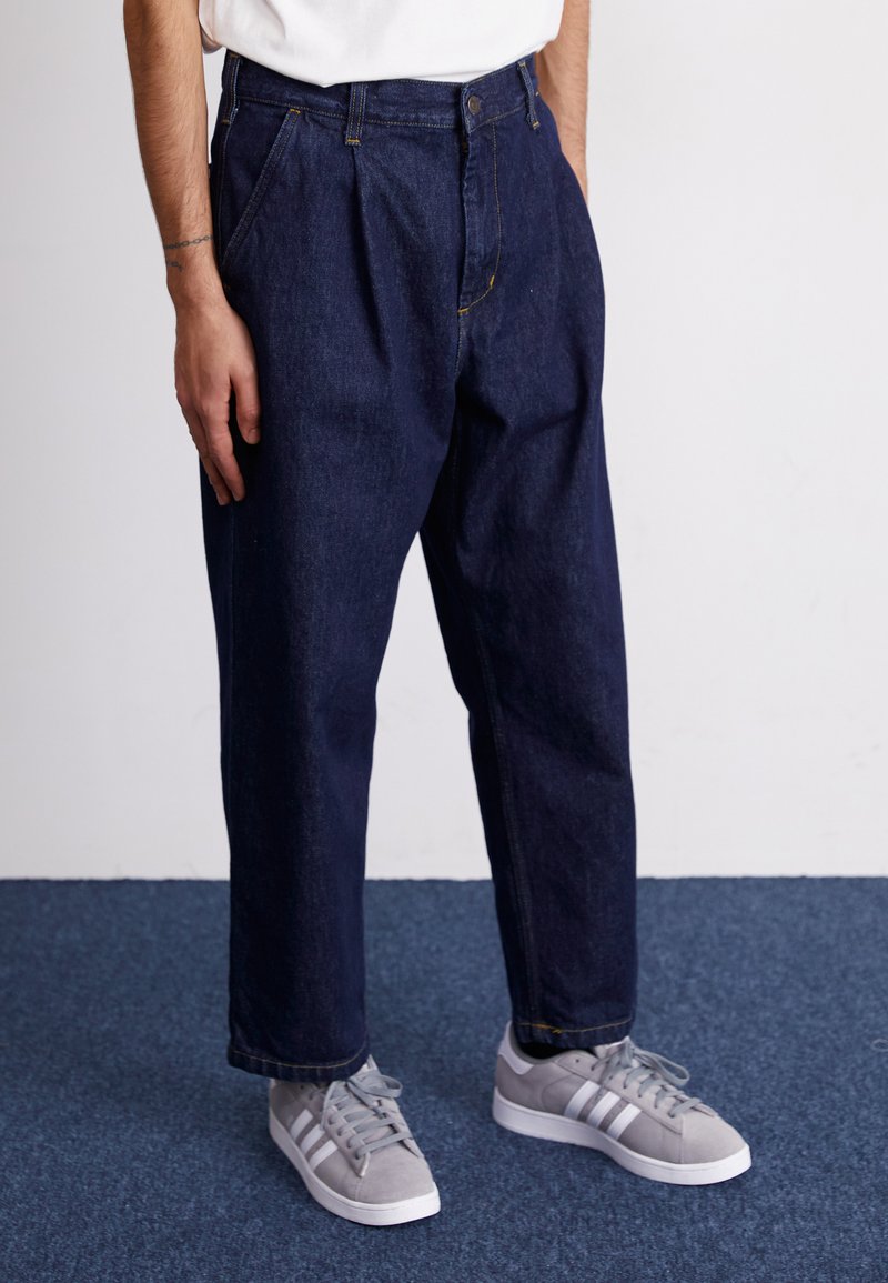 Donkerblauwe spijkerbroek met een relaxte pasvorm, voorzien van plooien, een knoopsluiting en oranje stiksels. Gekoesterd met grijze sneakers.