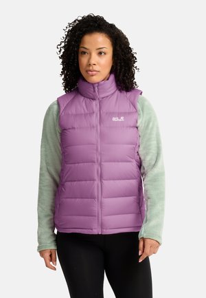 Frau mit lockigem Haar, die eine hellviolette ärmellose Daunenweste über einem grünen Fleece-Langarmshirt und schwarzen Hosen vor weißem Hintergrund trägt.