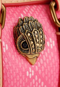 Decorazione a forma di gufo in oro e nero su uno sfondo rosa testurizzato con motivi circolari. Hardware include chiusure in tonalità oro.