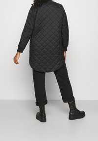 Vrouw in een lange zwarte gewatteerde jas, zwarte broek met omgeslagen mouwen en zwarte veterschoenen met plateauzool, staand tegen een effen lichte achtergrond.