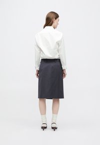 Sieviete ar muguru pret skatītāju, ģērbta baltā garroku blūzē, tumšā ceļos garā svārkā, baltas zeķes un brūnas papēžu kurpes uz vienkārša fona.