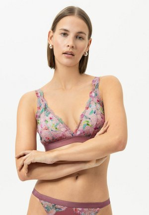 Jeune femme aux cheveux courts bruns portant un soutien-gorge en dentelle florale et une culotte assortie, debout, bras croisés sur fond blanc.