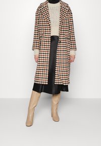 Karierter Mantel in Braun, Beige und Schwarz mit Reverskragen, großen Taschen und Strickärmeln, kombiniert mit einem schwarzen Satinenrock und beigefarbenen Stiefeln.