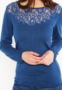 Sininen neule, jossa on pitsinen olkapääosa kukkakuviolla, pitkät hihat ja istuva malli. Pehmeä koostumus ja yhtenäinen väri kauttaaltaan.