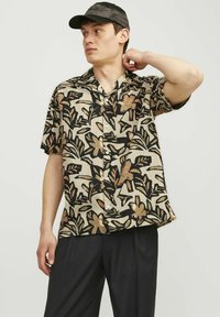 Chemise à manches courtes beige avec un motif floral noir, dotée d'un col et d'une fermeture à boutons, associée à un pantalon noir et une casquette.