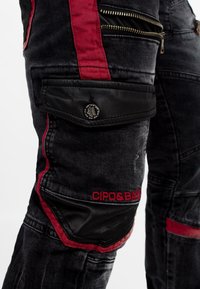 Pantalons cargo en denim noir avec des accents rouges et une poche en cuir, dotés de multiples zippers et d'un logo sur le côté. Détails de texture délavée.