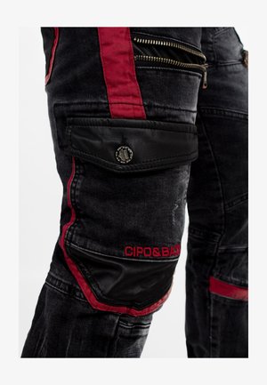 Pantalons cargo en denim noir avec des accents rouges et une poche en cuir, dotés de multiples zippers et d'un logo sur le côté. Détails de texture délavée.