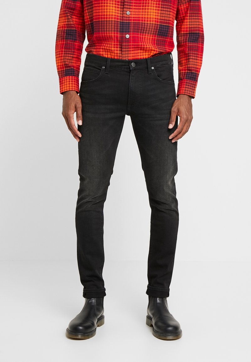 Homme portant un jean noir slim, des bottines noires et une chemise à manches longues à carreaux rouge et orange, debout devant un fond uni.