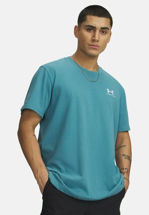 Mann mit kurzen dunklen Haaren trägt ein türkisfarbenes Under Armour T-Shirt, schwarze Hose, eine Kettenhalskette und hat Tätowierungen an den Unterarmen. Hände in den Taschen.