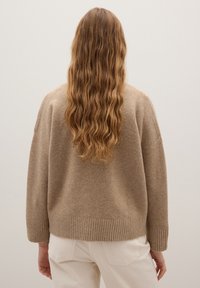 Beiger Wollpullover mit lockerem Schnitt, geripptem Saum und langen Ärmeln. Rückansicht zeigt weiche Textur und geraden Schnitt, getragen mit hellen Hosen.