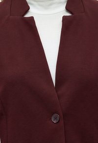 Gros plan d'un blazer texturé bordeaux avec un seul bouton sur un pull à col roulé blanc côtelé.