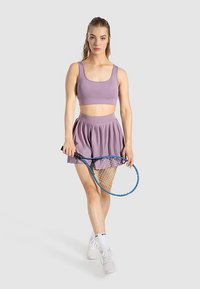 Top sportivo corto viola e gonna plissettata, con in mano una racchetta da tennis blu. Il modello indossa scarpe da ginnastica bianche e calzini alla caviglia.