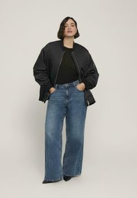 Giubbotto bomber nero con zip frontale, abbinato a jeans a vita alta blu con gamba larga. Il modello indossa un top nero e scarpe nere a punta.