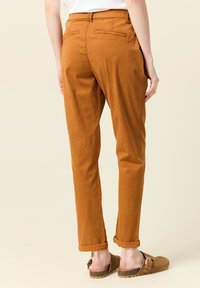 Brązowe bawełniane spodnie o luźnym kroju, zwężających się nogawkach i podwiniętych mankietach, z tylną kieszenią, noszone z brązowymi chodakami.