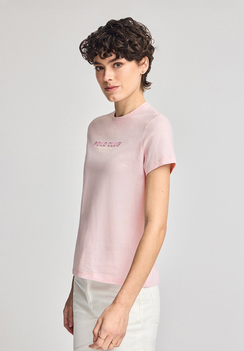 Ljusrosa t-shirt i mjuk bomull. Har rund halsringning, korta ärmar och texten "POLO CLUB" i skrivstil över bröstet.