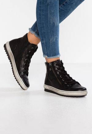 Czarne wysokie sneakersy z skóry z bocznym zamkiem i sznurowadłami noszone z niebieskimi dżinsami o postrzępionych nogawkach na białej podłodze.