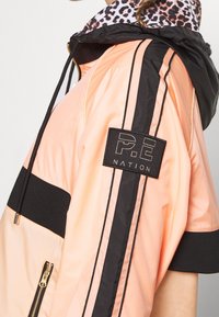 Chaqueta cortavientos melocotón y negra con capucha de leopardo, que incluye un bolsillo con cremallera, rayas negras y un parche de logo de P.E Nation.