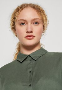Mørkegrøn skjorte med knaplukning og krave, lavet af et glat stof. Har sorte knapper og et enkelt design.