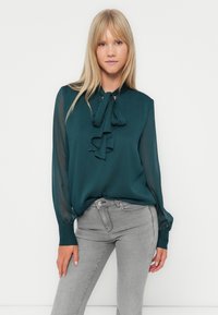 Blusa teal con colletto a fiocco, maniche lunghe trasparenti e tessuto testurizzato; abbinata a jeans grigi aderenti, che mettono in risalto una silhouette rilassata.