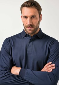 Tummansininen pitkähihaisessa polo-paidassa, jossa on teksturoitu kuvio, nappikaulus ja muotoilu, jota käyttää mies, joka on ristijättänyt kädet.