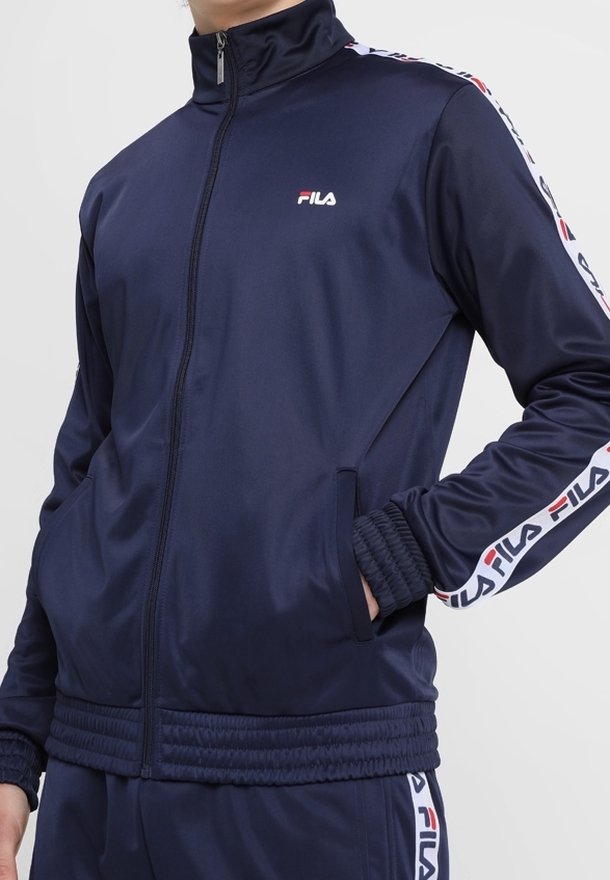 Mann trägt marineblauen FILA-Trainingsanzug mit weißen und roten Logostreifen an Ärmeln und Hosen, Hände in den Jackentaschen.