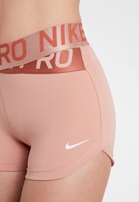 Detailní záběr osoby v broskvově zbarvených atletických šortkách Nike Pro s dvojitým páskem, na kterém jsou loga "NIKE" a "PRO".
