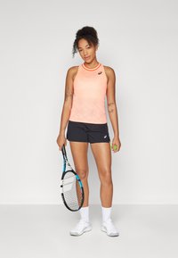 Tennis outfit featuring a bright orange sleeveless top with diamond pattern and black shorts. Model holds a tennis racket and ball. White shoes.