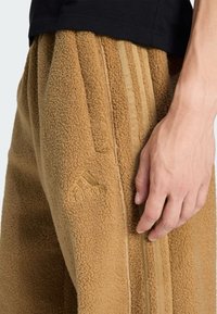 Braune Fleece-Hosen mit strukturiertem Finish, versehen mit vertikalen Streifen und einem gestickten Logo. Hand ruht auf der Seitentasche.