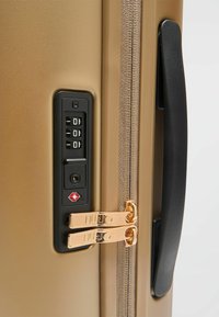 Valise rigide en or avec une finition lisse et texturée. Comprend un verrou à combinaison, une poignée noire et des fermetures éclair métalliques doubles avec le logo de la marque.