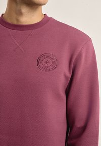 Sudadera color burdeos con tejido texturizado, cuello redondo y un logotipo circular embellecido en el pecho. Diseño suave y casual ideal para superponer.