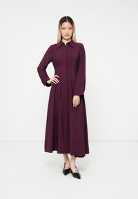 Junge Frau, die ein langärmliges, kragenloses, knöchellanges Kleid in Pflaumenfarbe mit Falten trägt, steht mit einer Hand an der Hüfte und trägt schwarze spitze Schuhe.