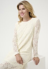 Krajková šaty v krémovej farbe, s dlhými krajkovými rukávmi s kvetinovými vzormi, jemnou textúrou a okrúhlym výstrihom. Hladký strih po celom obvode.