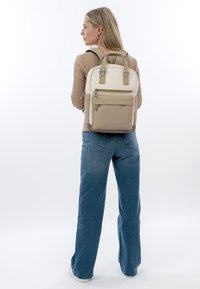 Femme portant un sac à dos beige et crème, un pull côtelé beige, un jean bleu et des chaussures blanches, debout devant un fond blanc uni.