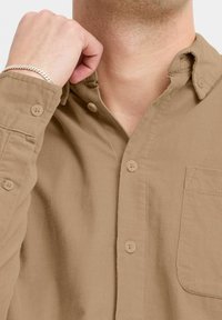 Camisa de pana marrón claro con cuello abotonado, bolsillo delantero y mangas largas. Presenta una textura acanalada y botones a juego.