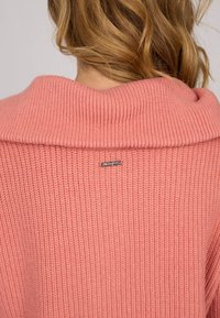 Pull en tricot de couleur rose doux, avec un col large et replié. Texture côtelée prononcée et un petit logo en métal près de l'encolure.