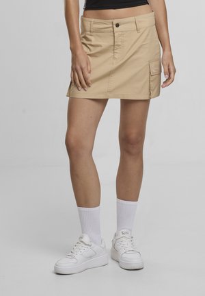 Donna che indossa una minigonna cargo beige, calzini bianchi e scarpe da ginnastica bianche, in piedi contro uno sfondo grigio uniforme.