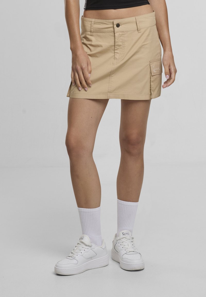 Femme portant une mini-jupe cargo beige, des chaussettes blanches et des baskets blanches, debout devant un fond gris uni.