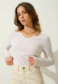 Vit långärmad tröja med vågig halsringning och fyra små knappar på vänster sida. Mjuk textur, bärs med ljusbeiga jeans.
