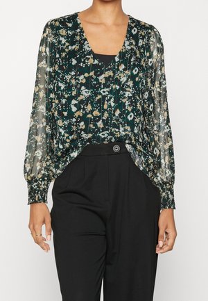 Vrouw die een doorschijnende blouse met groene bloemenprint draagt, met lange pofmouwen en zwarte, hoog uitgesneden broek met knoopsluiting.