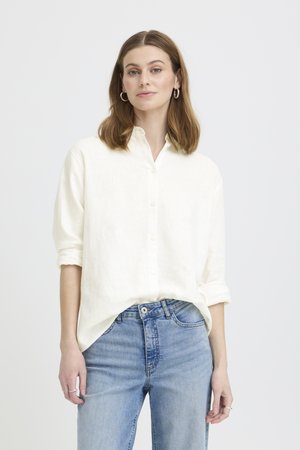 Jeune femme aux cheveux bruns mi-longs, portant une chemise blanche à boutons avec manches retroussées et un jean bleu clair, debout devant un fond blanc.
